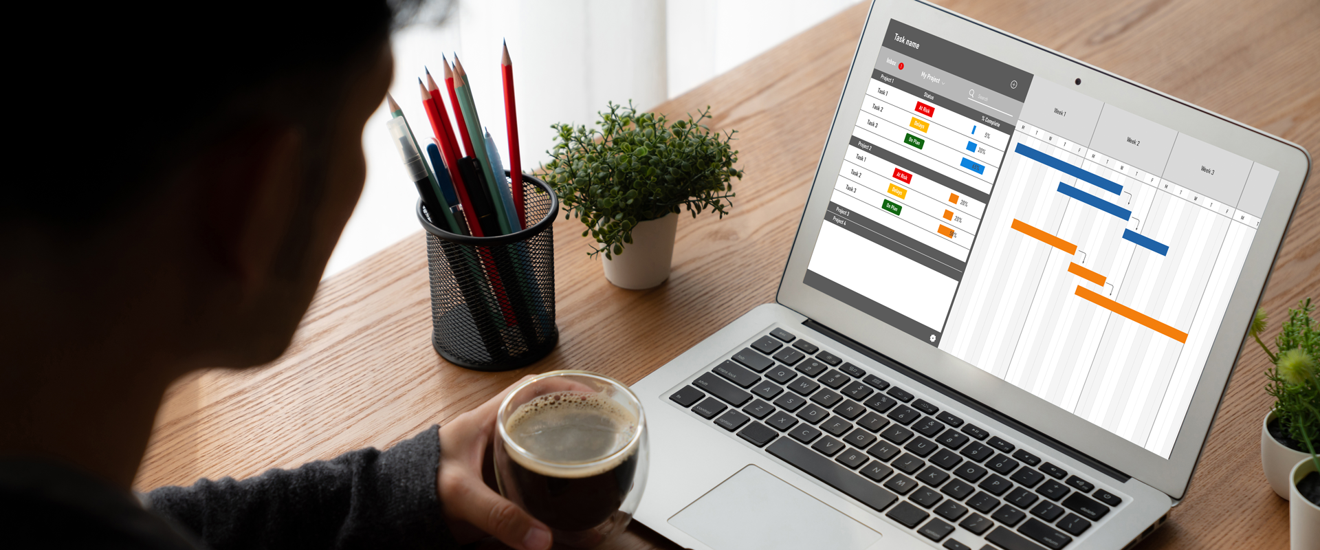 Person holding coffee while working on a laptop with a project management software interface and office supplies.
